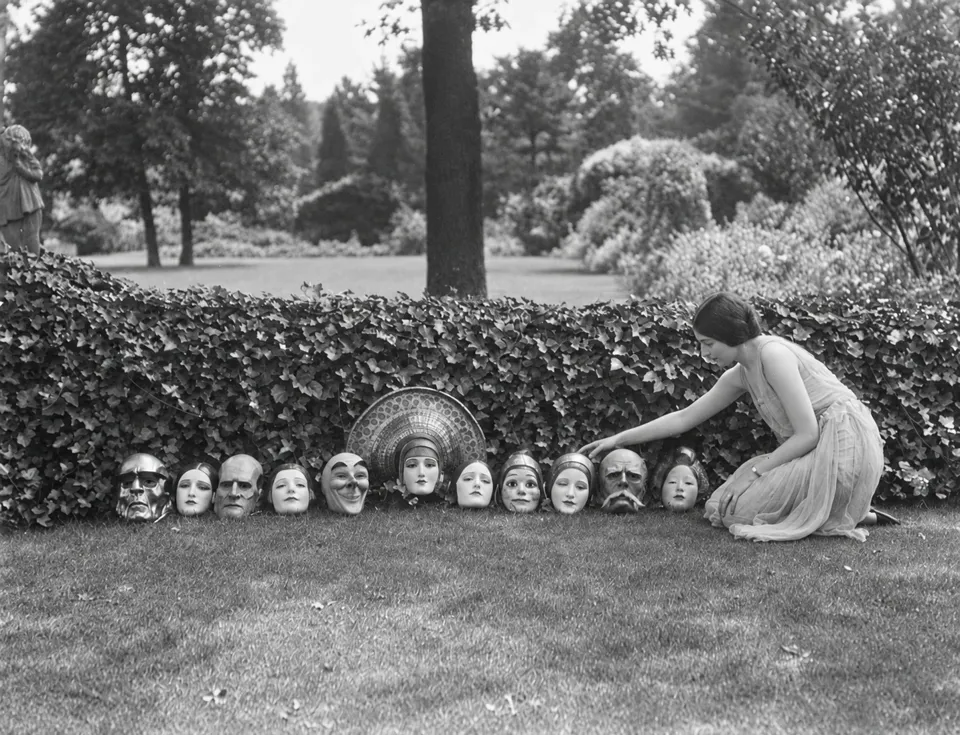 A woman kneels beside a display of W. T. Benda’s theatrical masks in Arnold Genthe’s photograph.