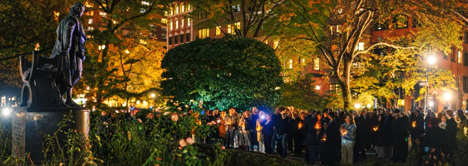 The Booth statue at left overlooks a nighttime gathering of people carrying glowing lanterns beneath autumn trees.
