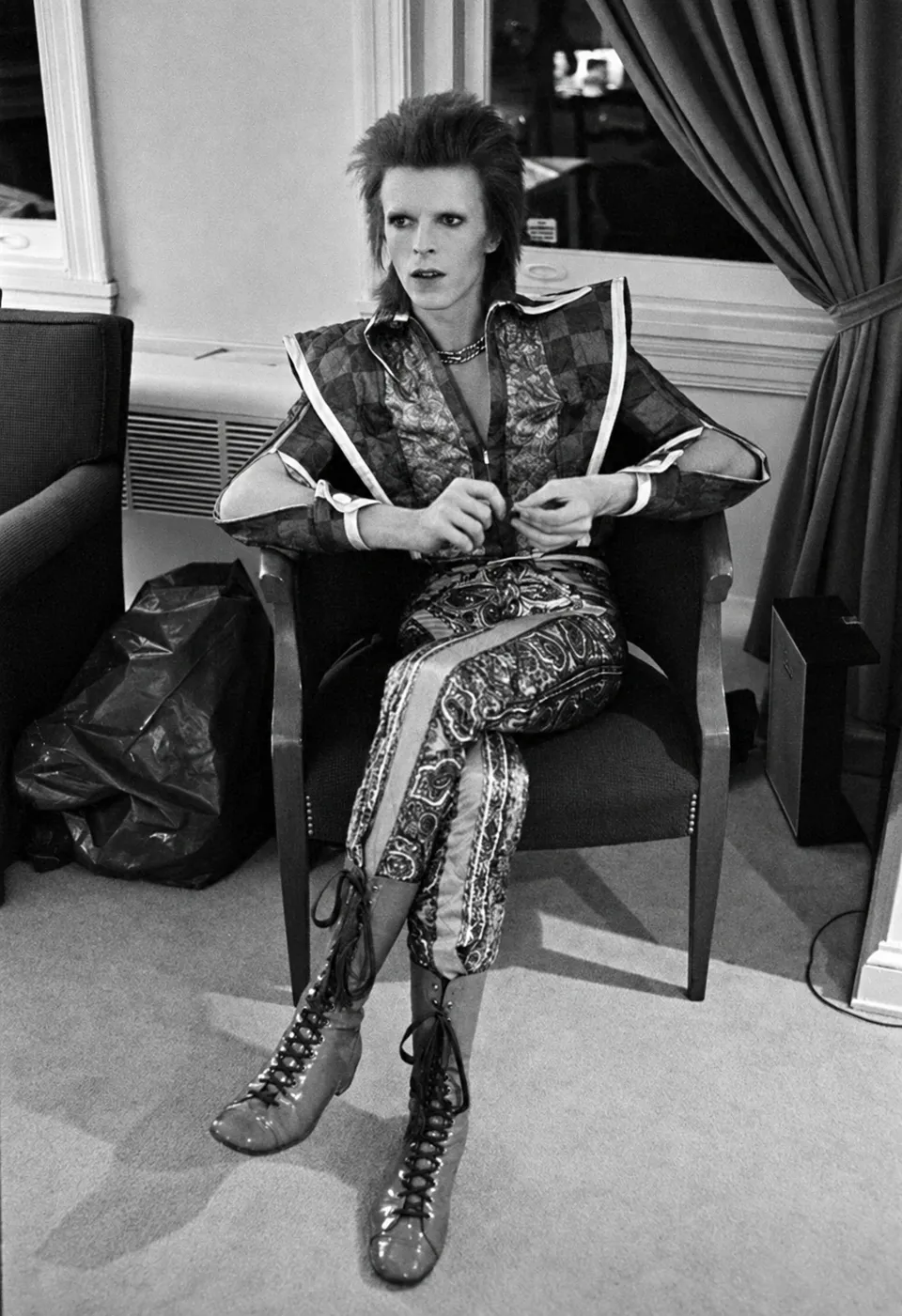 David Bowie seated in an armchair, wearing an ornate outfit and high lace-up boots.
