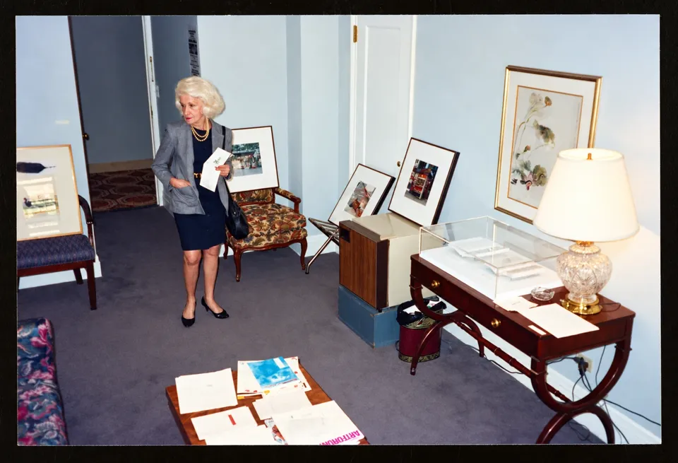 A visitor in a hotel guest room used as an exhibition space, with framed artworks propped against furniture and walls.