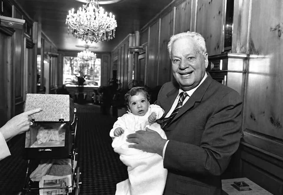 Herbert Weissberg holding baby Max in a paneled hotel corridor, beside a baby carriage.