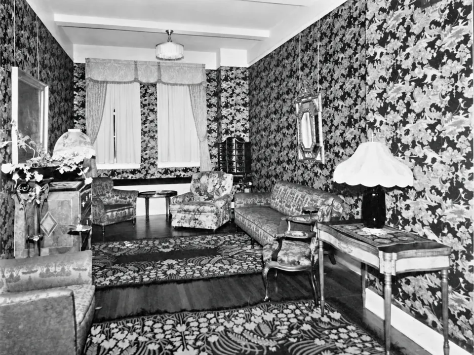 A hotel sitting room with floral wallpaper, upholstered chairs, a sofa and a table lamp.