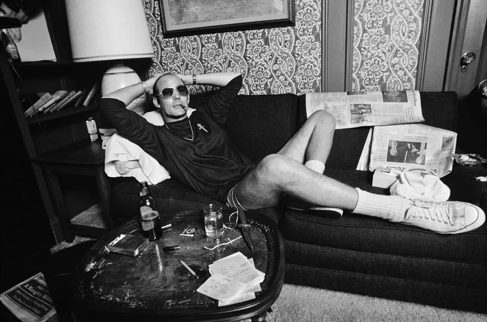 Hunter S. Thompson reclining on a sofa at the Gramercy Park Hotel in 1977, with newspapers and a table in the foreground.