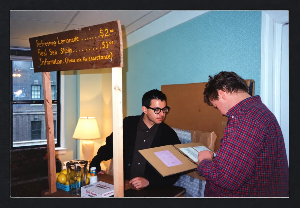 Mark Dion behind a lemonade stand in a hotel room, with a visitor looking through a book.