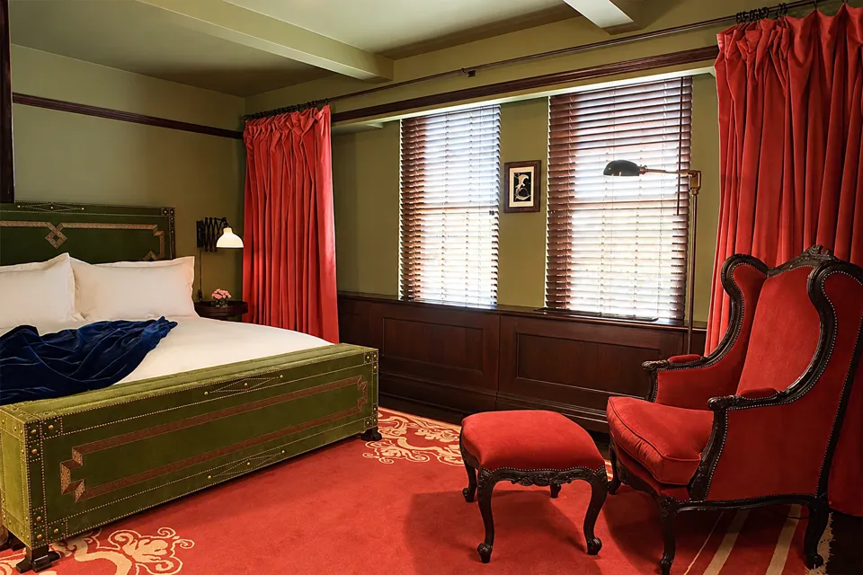 A green upholstered bed, red armchair and curtains, patterned carpet, and tall windows in a hotel guest room.