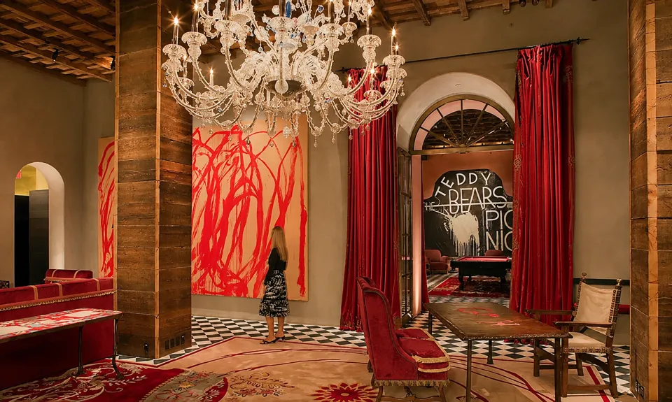 A crystal chandelier above the lobby’s wood columns, large paintings, arched doorway, red curtains, and checkerboard floor.