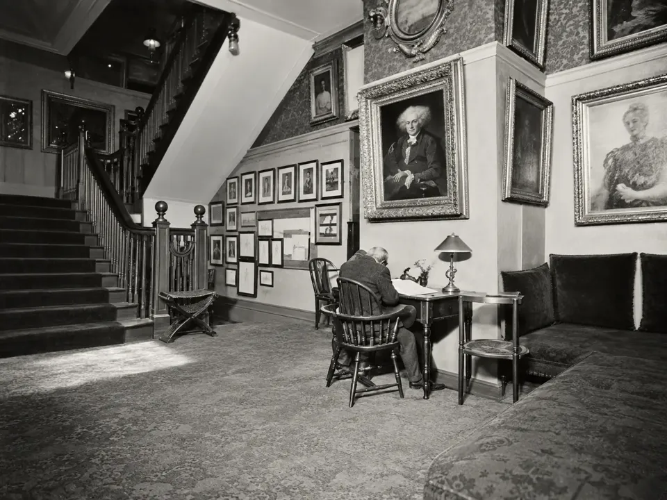 The second-floor hall of The Players in a 1905 photograph, with the staircase, framed pictures and a seated reader.