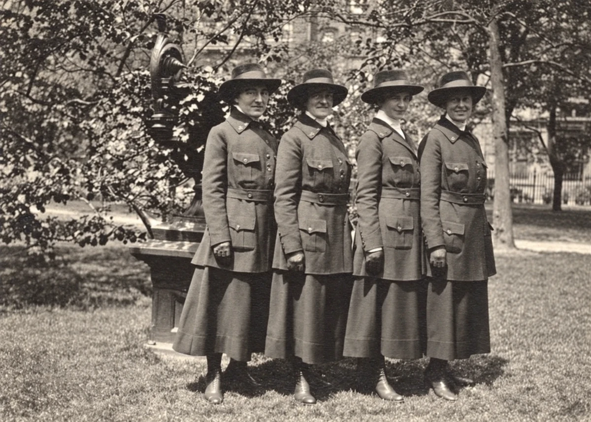 Four nurses in belted uniform jackets, long skirts, and brimmed hats stand together among the trees of Gramercy Park in May 1918.