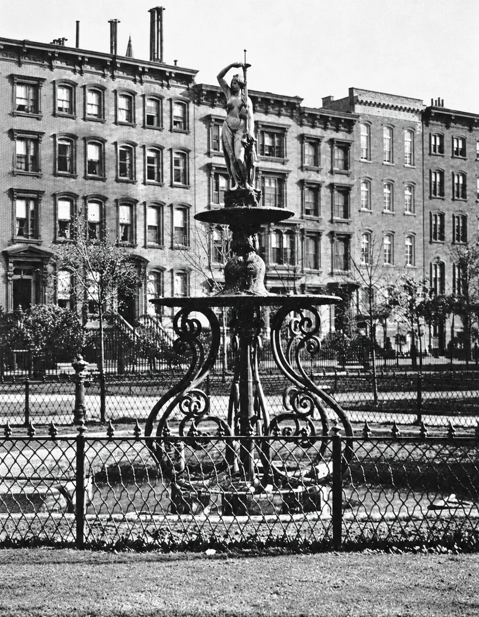 A historic photograph of a tall, tiered fountain topped by a female figure amid trees and garden paths.
