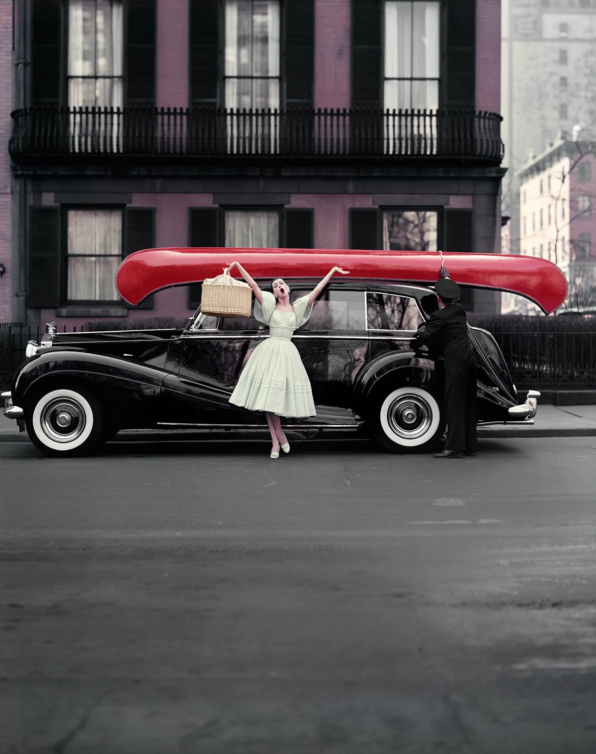 Barbara Mullen raises her arms beside a black car carrying a bright red canoe, with shuttered brick houses behind her.