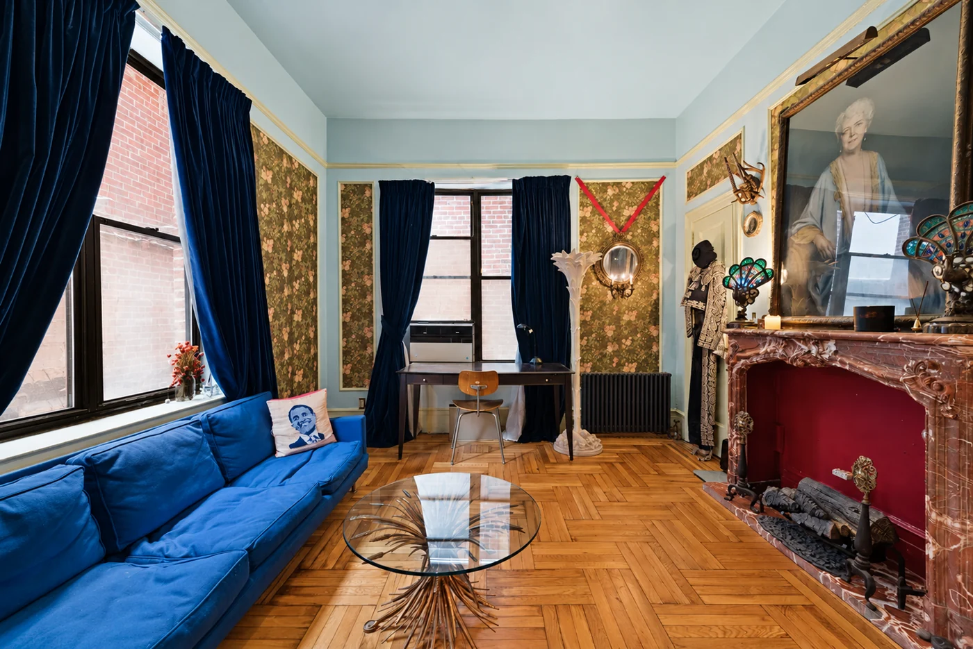 A blue sofa faces a fireplace in a compact studio with scenic wallpaper and a writing desk by the windows.