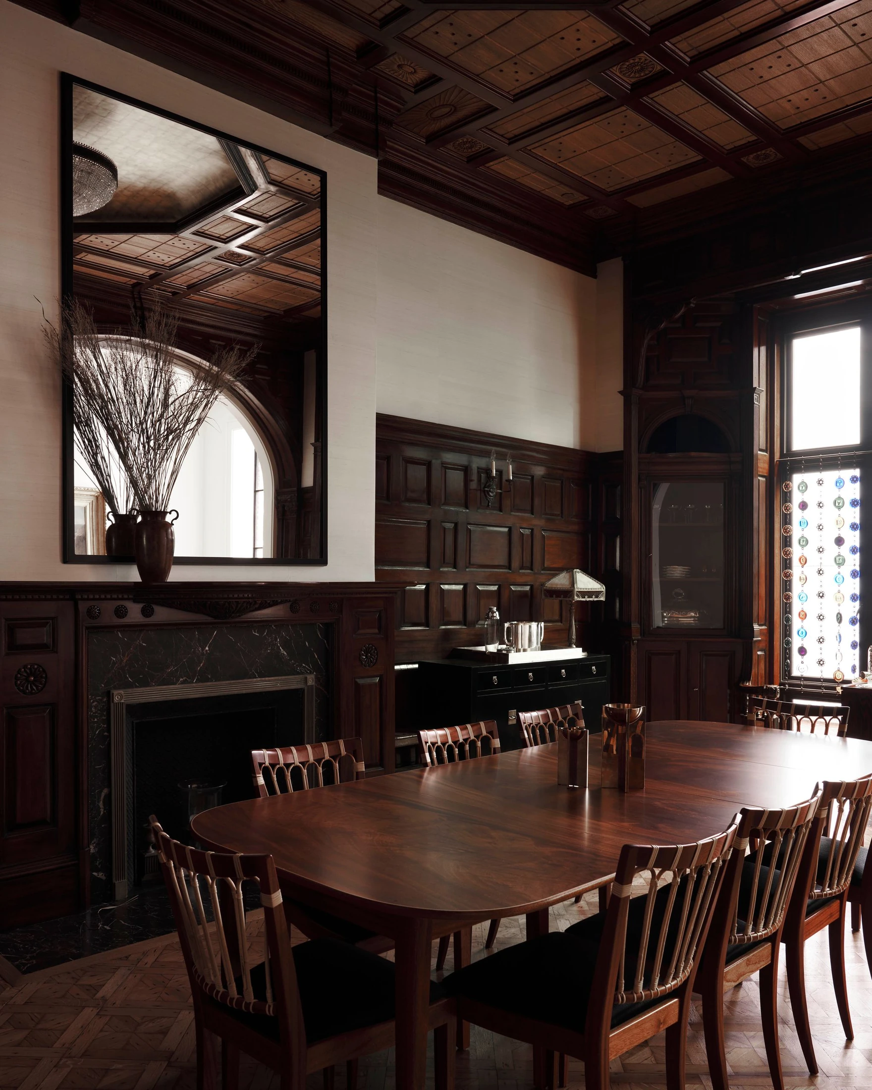 The restored dark wood salon with coffered ceiling, fireplace, dining table and tall window.