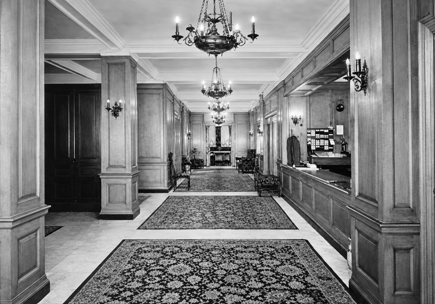 Historic view of the Gramercy Park Hotel lobby, with paneled columns, chandeliers, patterned rugs and the reception desk on the right.