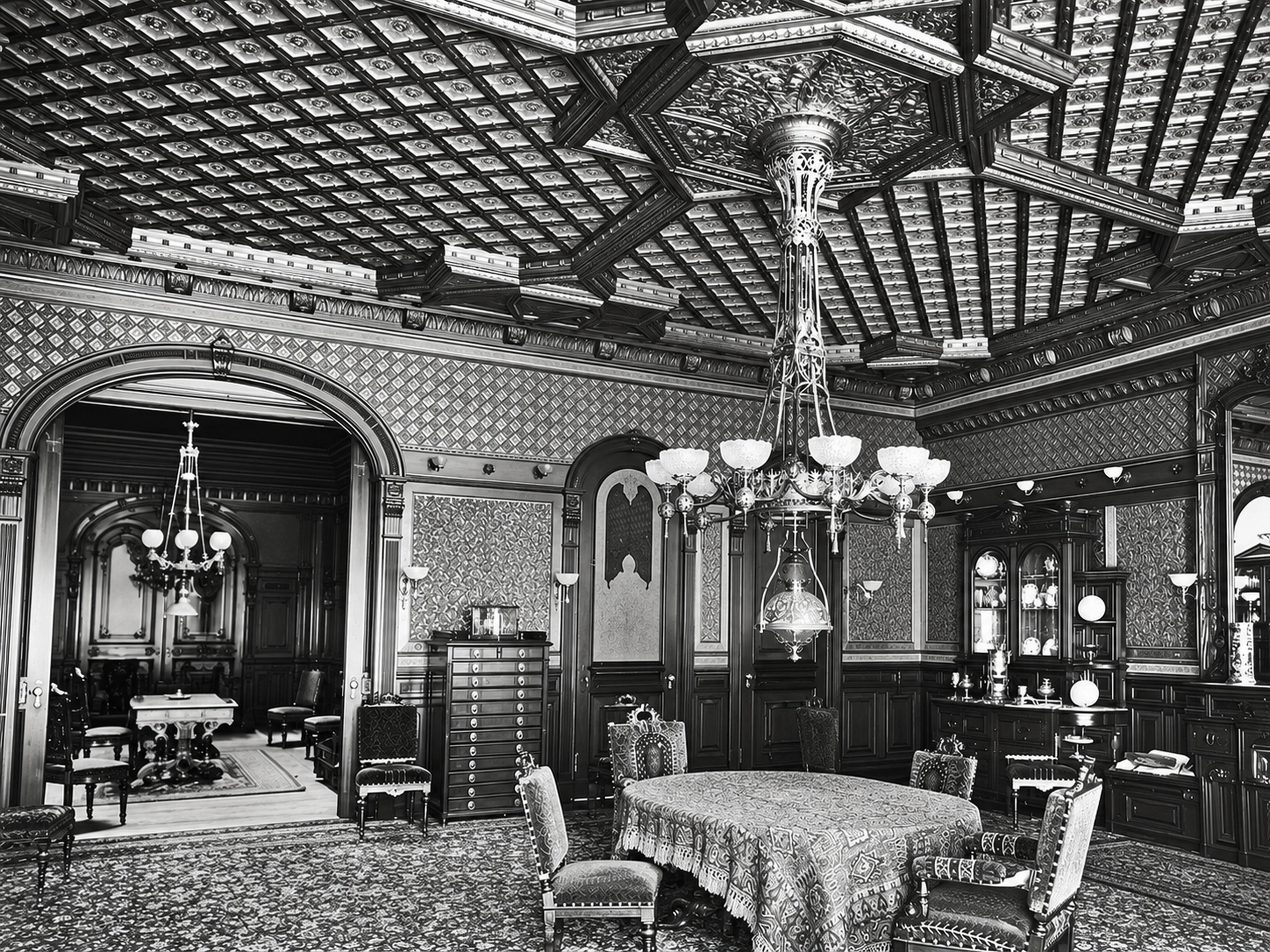 An archival photograph of an elaborately decorated room with a tiled ceiling, chandeliers, wood paneling and a round dining table.