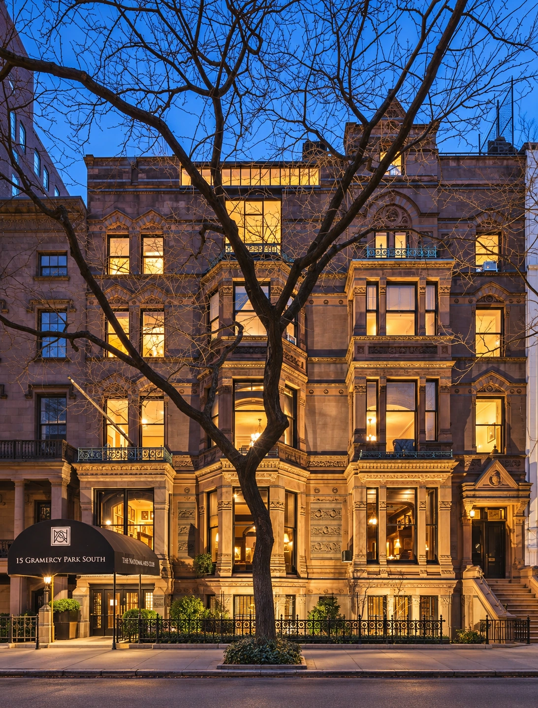 The full facade of the Tilden Mansion at dusk, with warm light shining from the windows and bare tree branches in front.