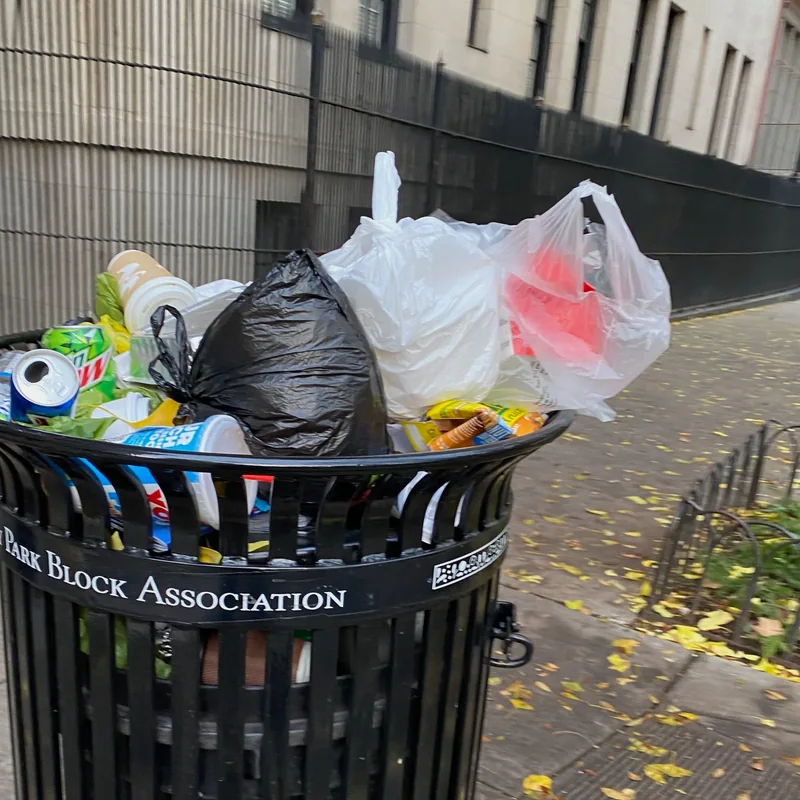 An overflowing litter basket beside a building fence.