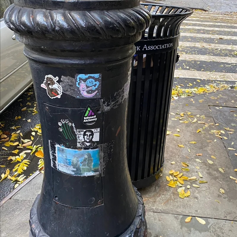 Stickers cover a black lamppost base beside a litter basket.