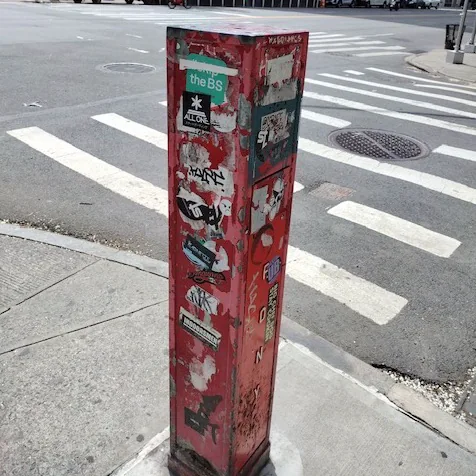 Posters and stickers cover a red street box beside a crosswalk.