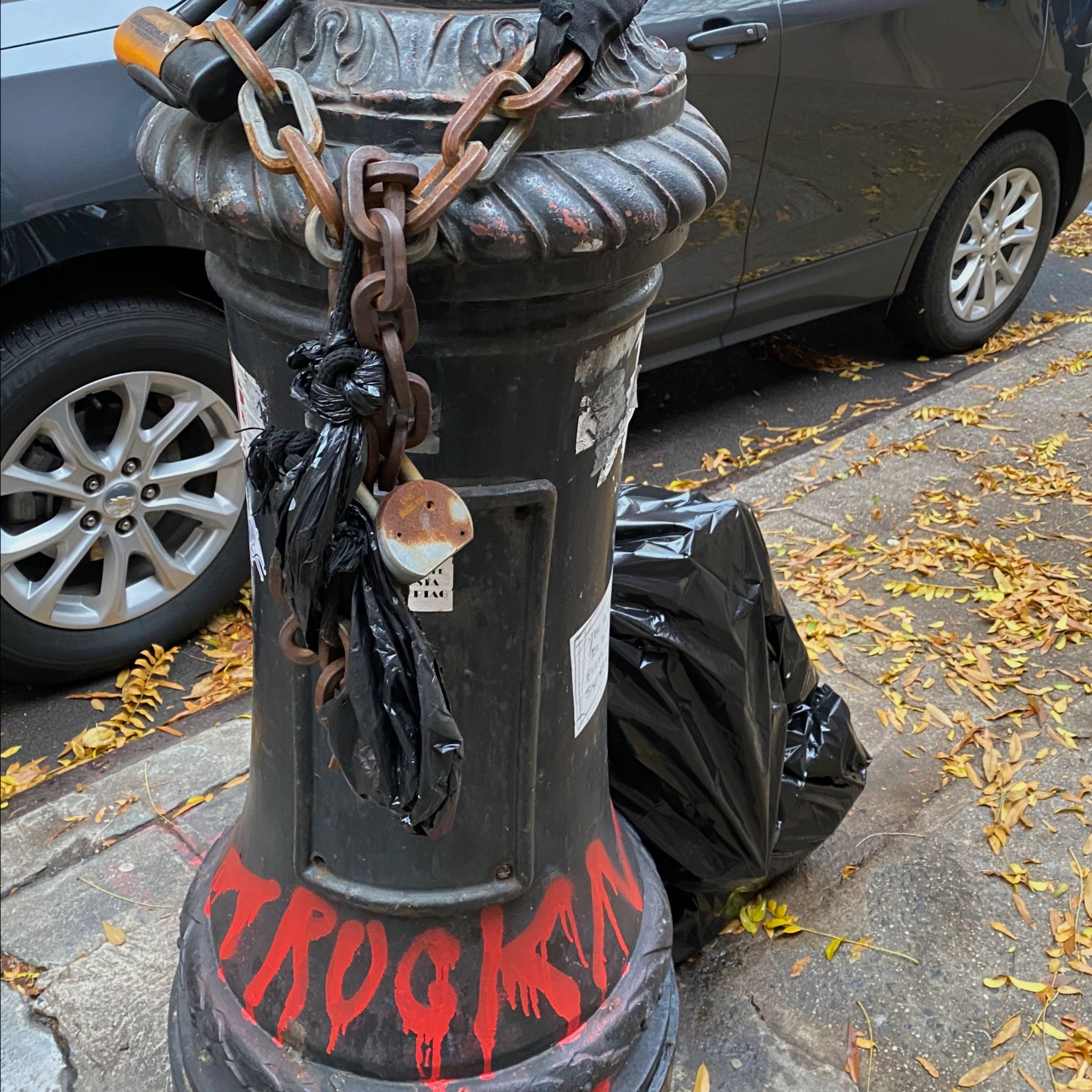 The base of a black lamppost has graffiti, stickers and a chain attached before maintenance.