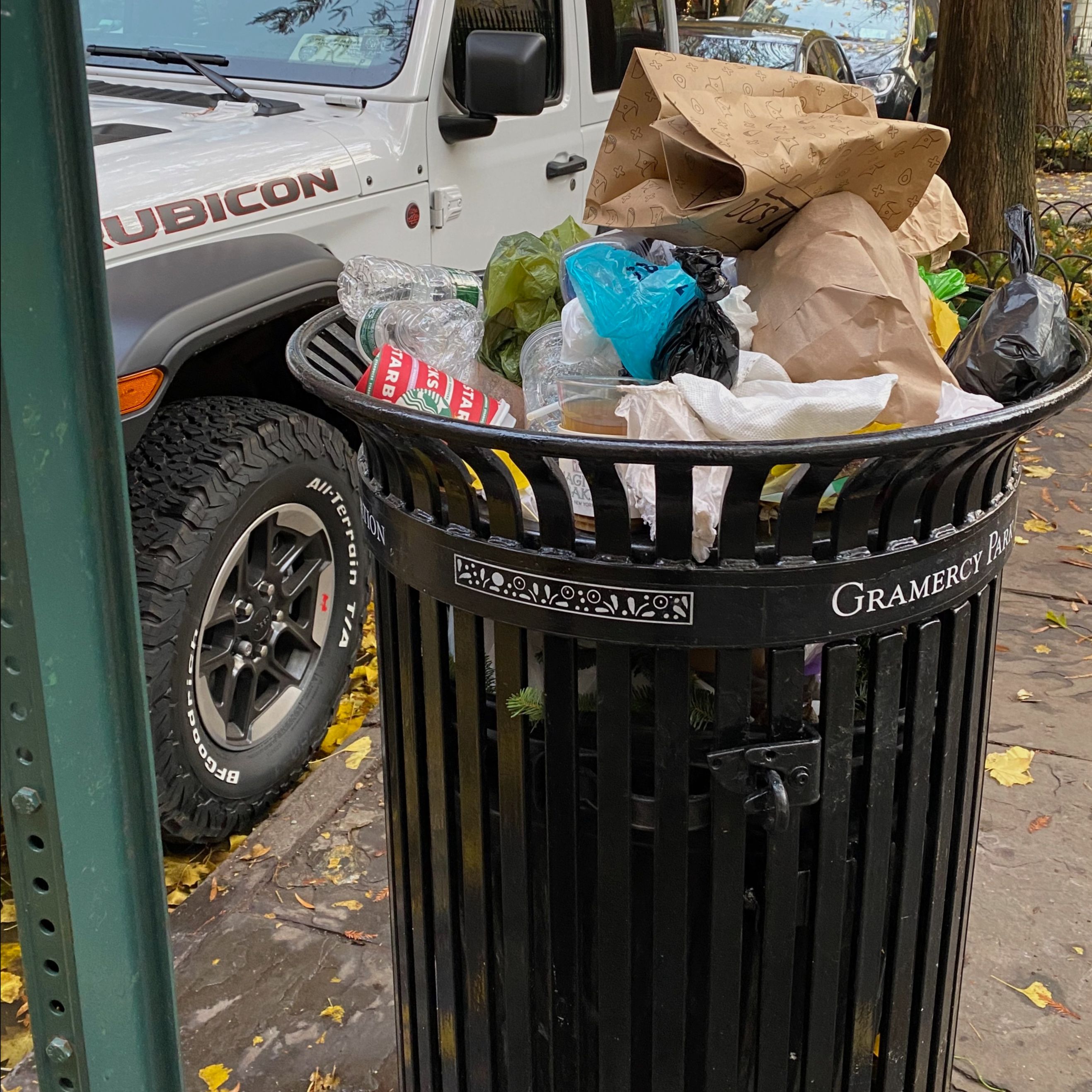A sidewalk trash receptacle is overflowing with paper bags and other refuse.