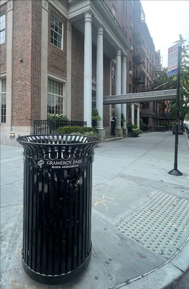 A black the Block Association litter receptacle stands on the sidewalk outside a building with tall white columns.