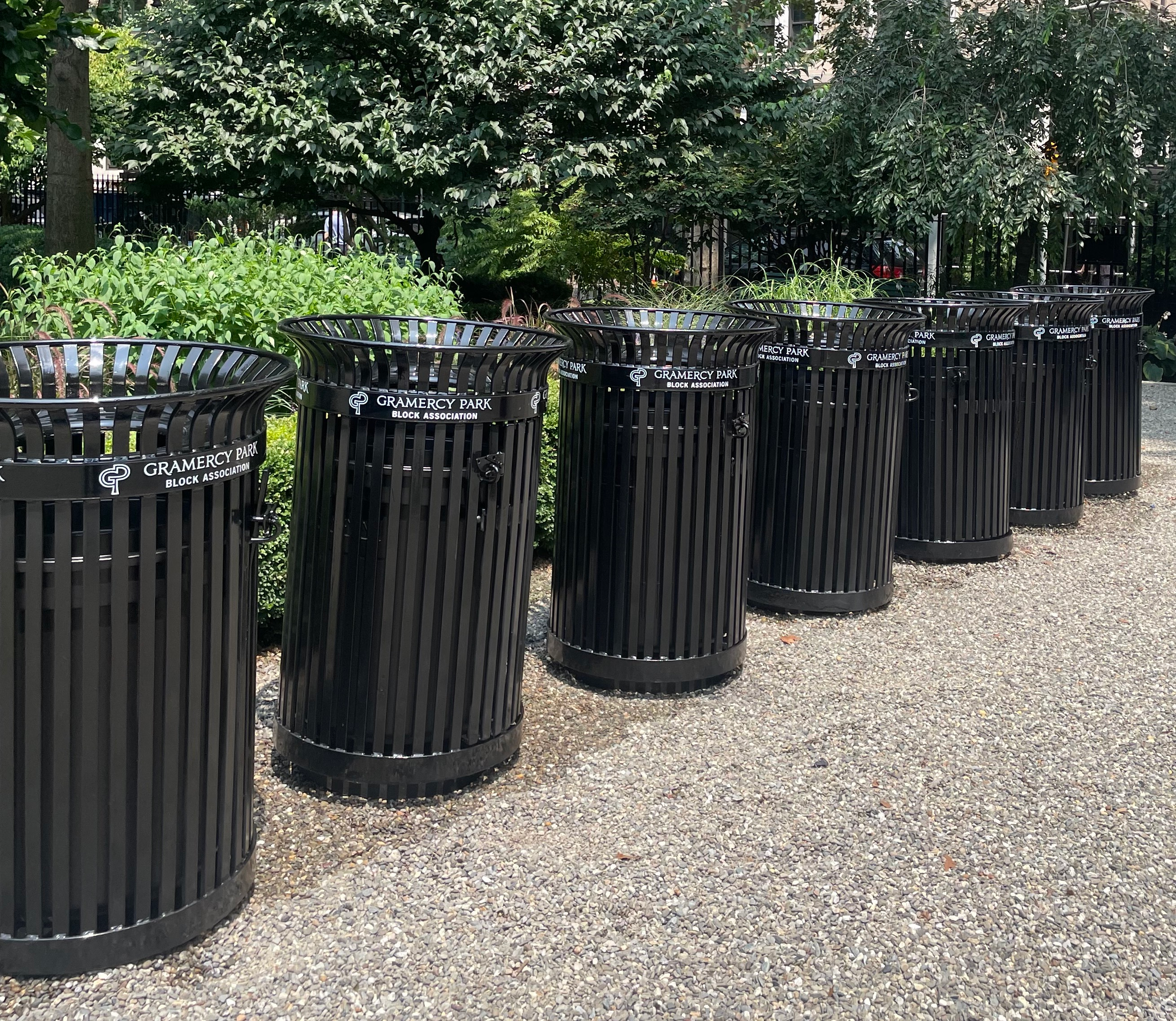A row of black metal litter receptacles bearing the Block Association name stands along a gravel path.