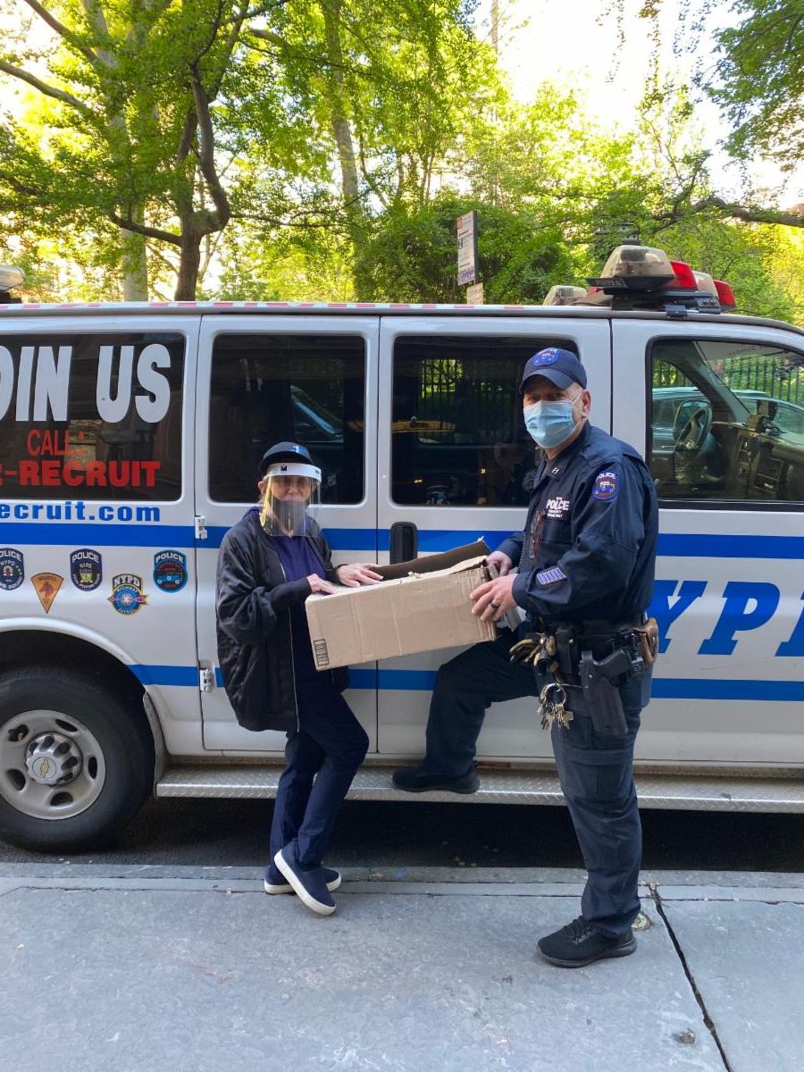 Two masked people hold a cardboard box beside an NYPD van on a tree-lined street.