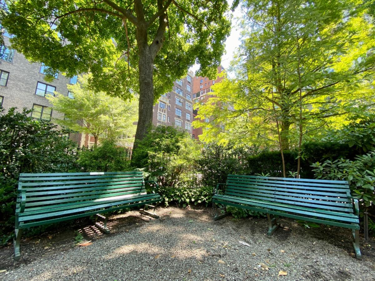 Two green benches meet at a shaded corner surrounded by shrubs and trees inside Gramercy Park.