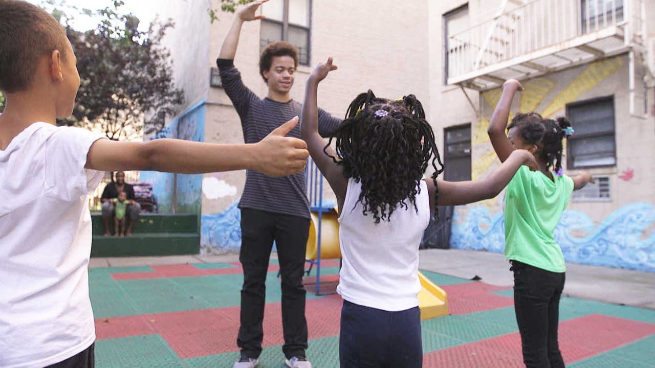Steven Melendez leads an outdoor dance exercise with Project LIFT students, their arms raised outward.