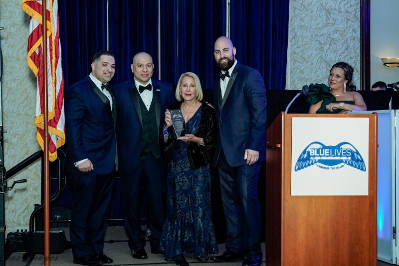 Arlene Harrison holds an award beside three men at a Blue Lives Matter NYC presentation, with a woman at the podium.