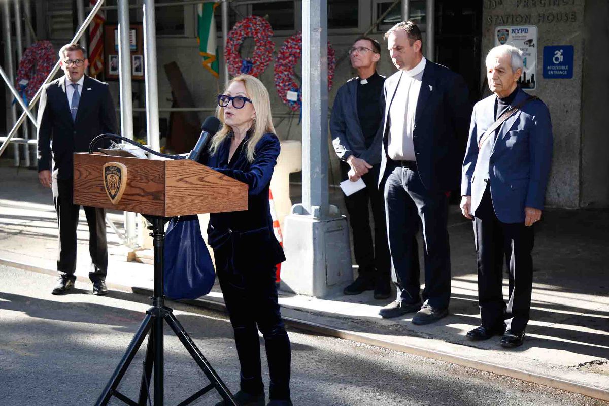A speaker stands at a wooden podium outside the 13th Precinct with other attendees nearby.