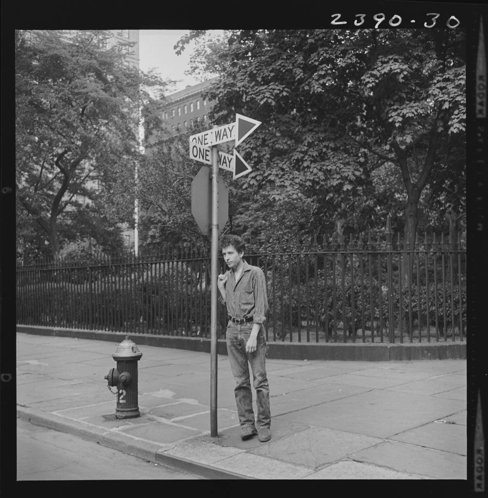 Bob Dylan leaning against a street sign beneath two one-way arrows beside Gramercy Park, 1963.