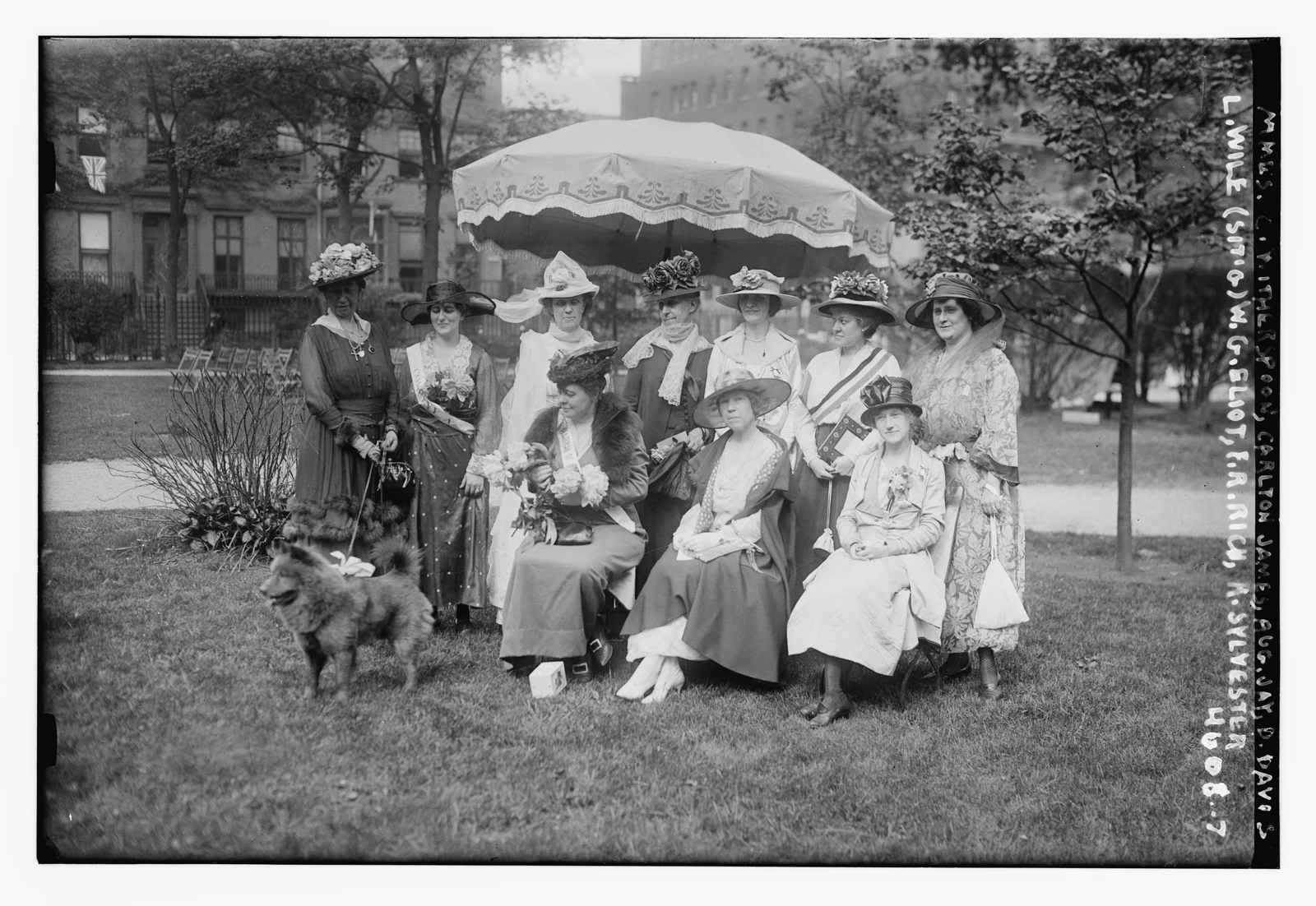 Women seated and standing beneath a fringed parasol, with a dog in front.