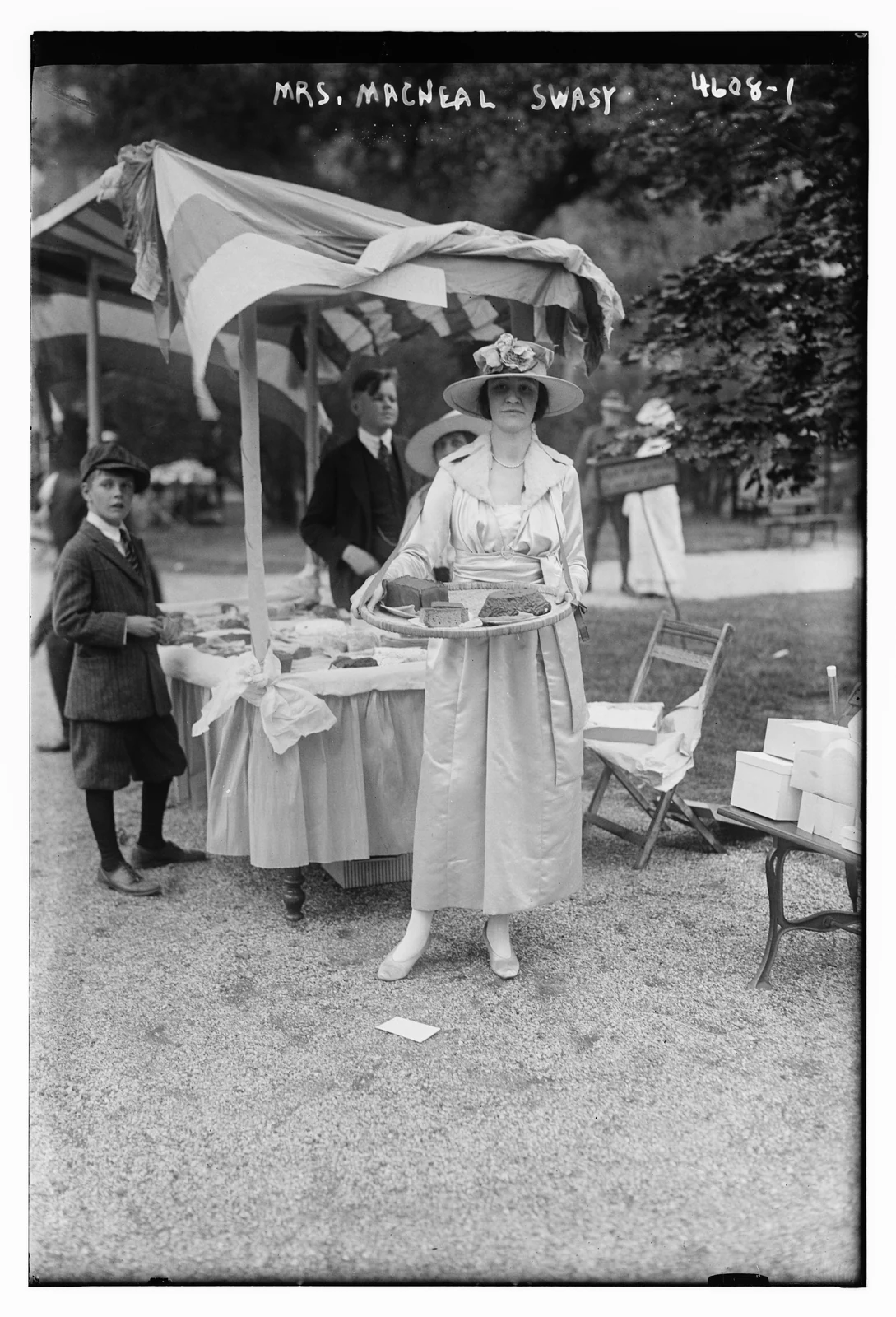 A woman holds a tray beside a refreshment stall, with a boy and a man nearby.