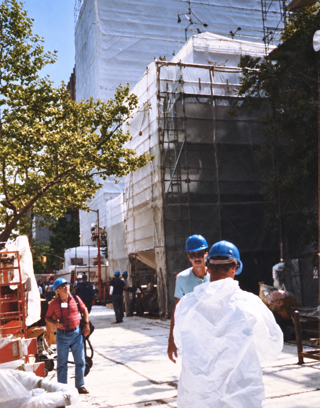 Workers in hard hats stand beside scaffolding and a building enclosed in white sheeting.