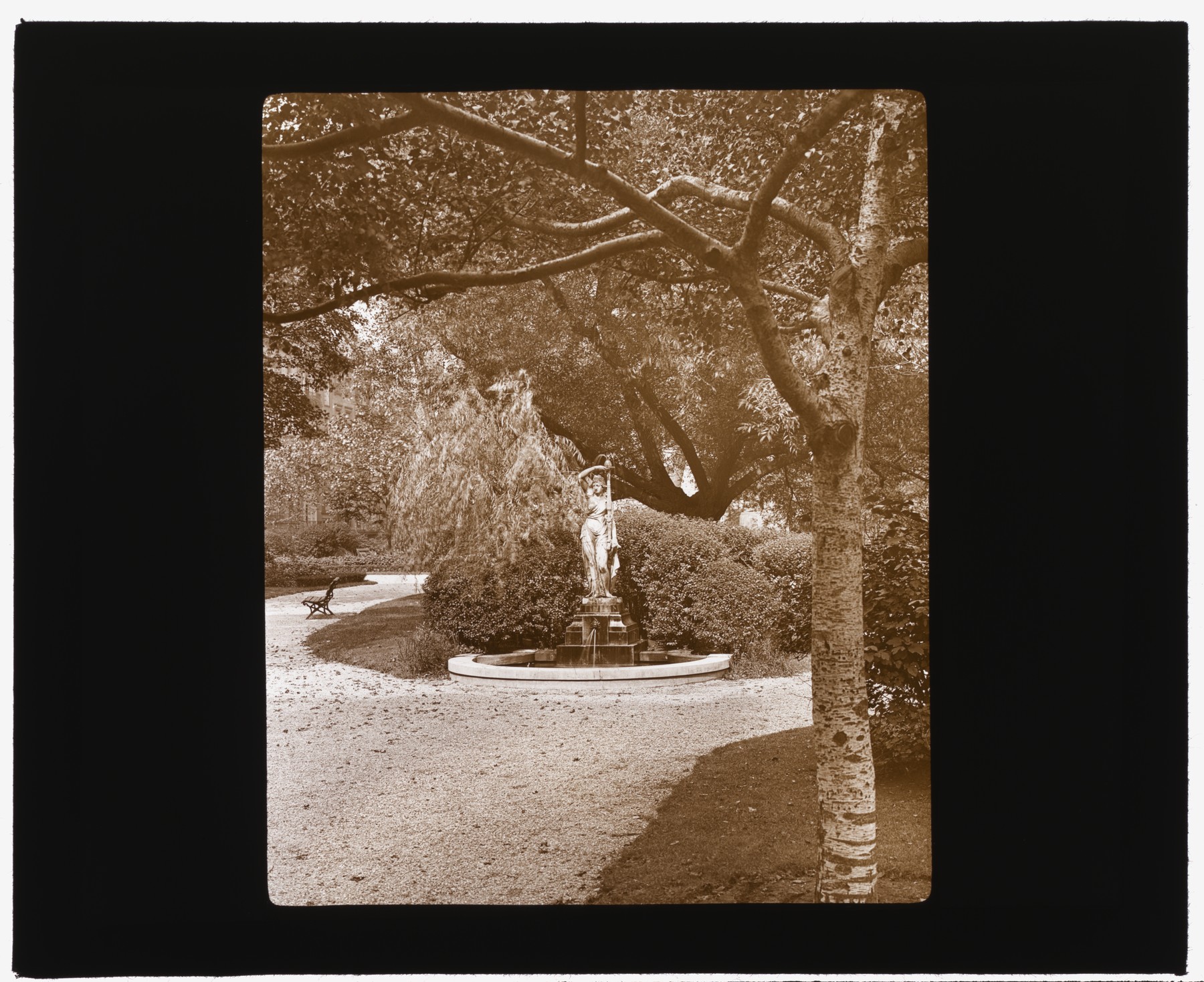 A female figure rises above a round fountain basin beneath trees in Gramercy Park, in a sepia lantern slide from about 1922.