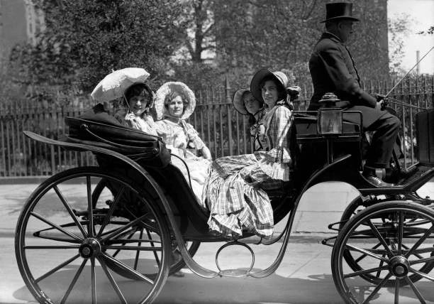 Three women in old-fashioned dresses sit in an open carriage, with a driver on the raised seat.