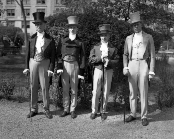 Four men in top hats, tailcoats and light trousers stand together on the lawn.