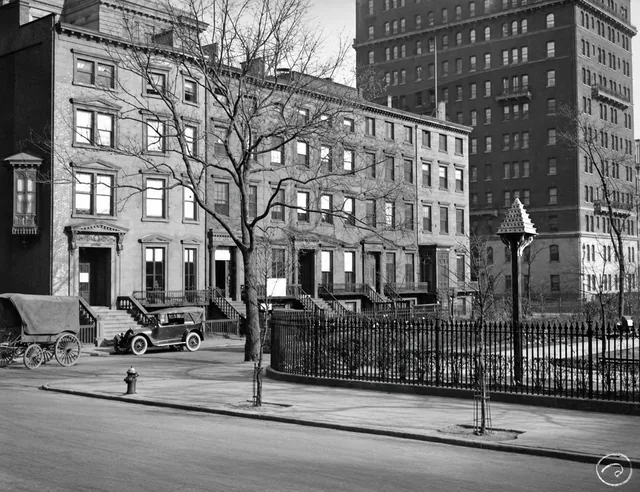 Row houses on Gramercy Park North beside a tall apartment building, with an automobile, carriage and the Park’s iron fence in the foreground.
