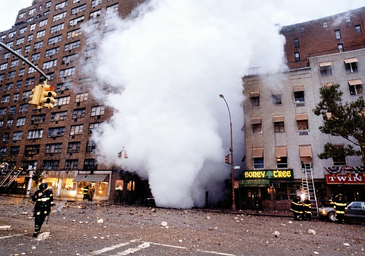 A plume of steam rises between buildings on Gramercy Park South as firefighters stand in a street covered with debris.