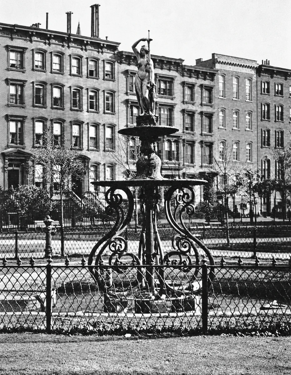 A historic photograph of a tall, tiered fountain topped by a female figure amid trees and garden paths.