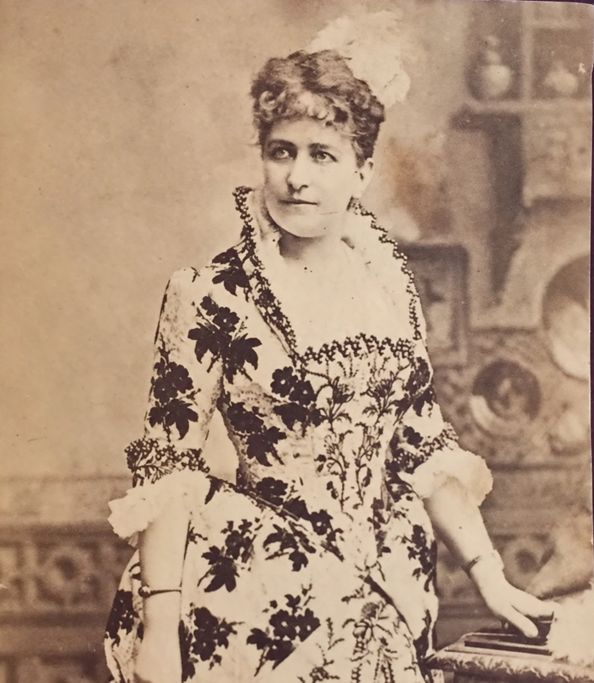 Emma Thursby beside a piano, wearing a floral embroidered gown and a feather in her hair.