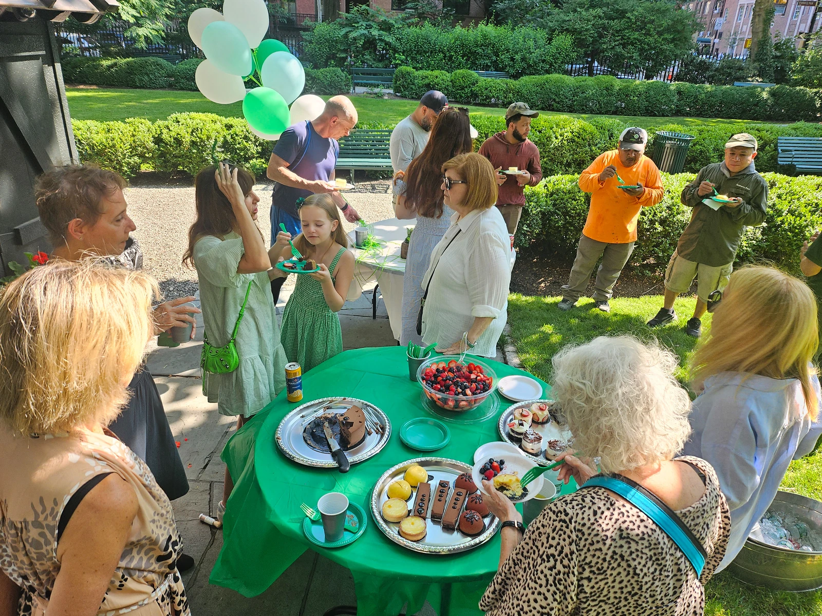 Neighbors gather around a table of birthday treats beneath green and white balloons in the Park.