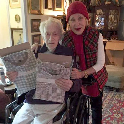 A visitor stands beside an older person in a wheelchair who holds two gift bags.