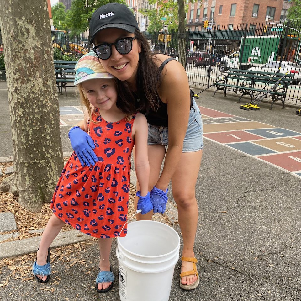 An adult and child wearing cleaning gloves stand beside a bucket in a playground.