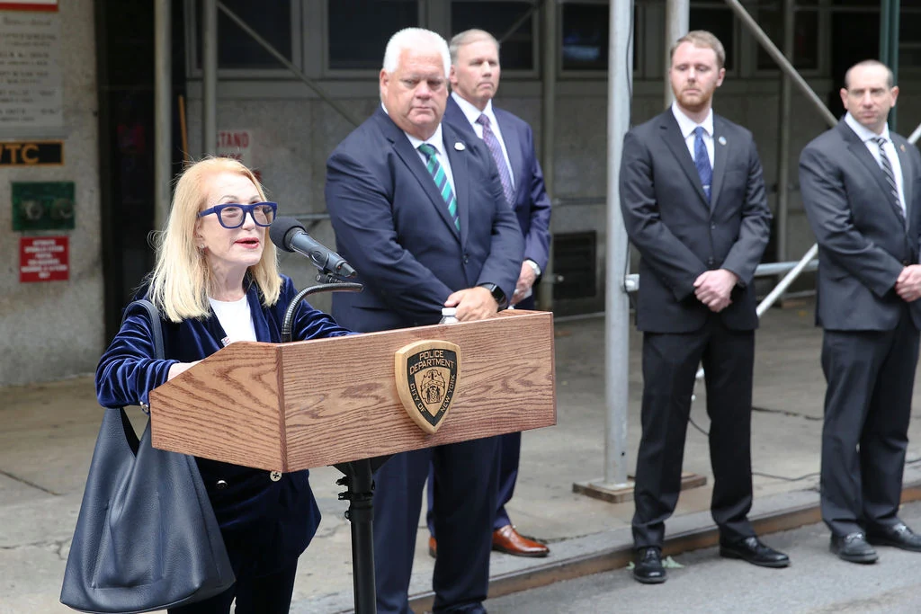 A woman speaks at a podium outside the precinct with attendees standing behind her.