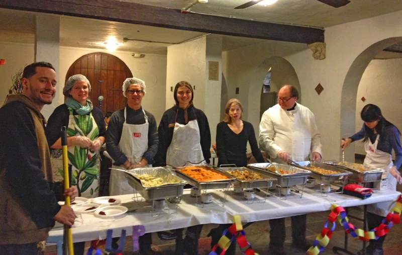 A group of volunteers stands behind trays of food in a church dining room.
