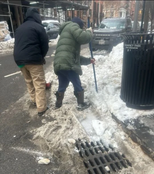 Two workers use shovels to clear slush beside a street drain.